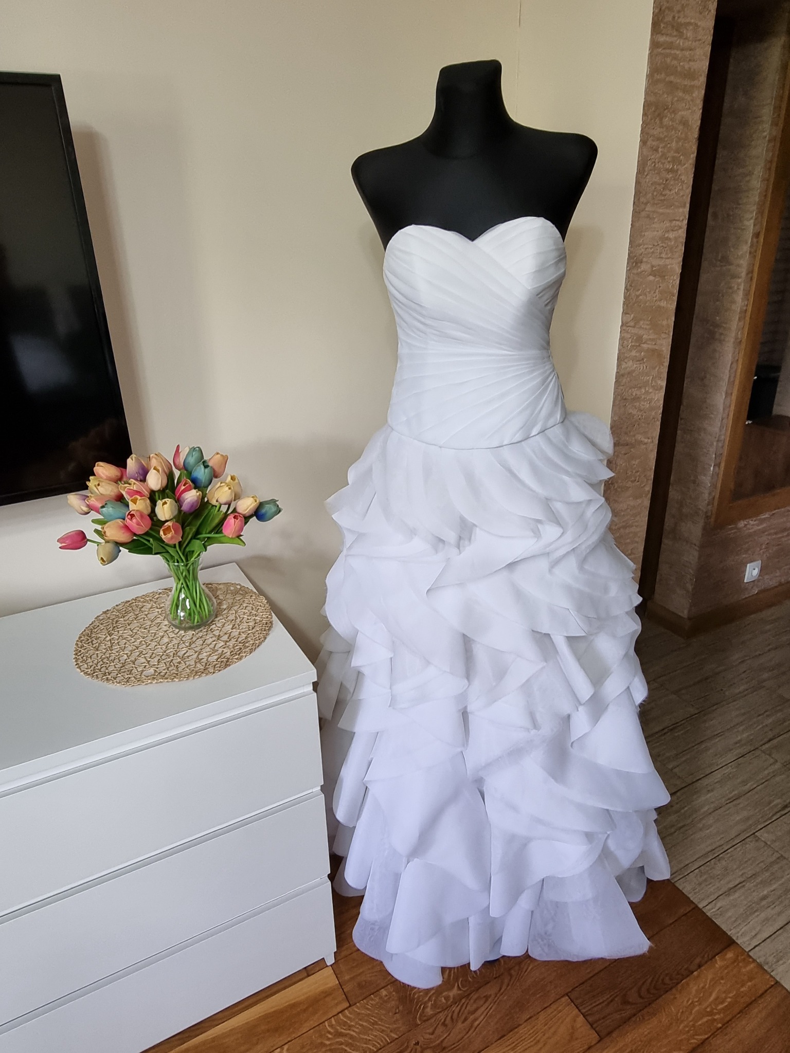 Elegancka suknia ślubna bez ramiączek, z falbanami na dole, prezentowana na czarnym manekinie obok białej komody z bukietem kolorowych tulipanów.