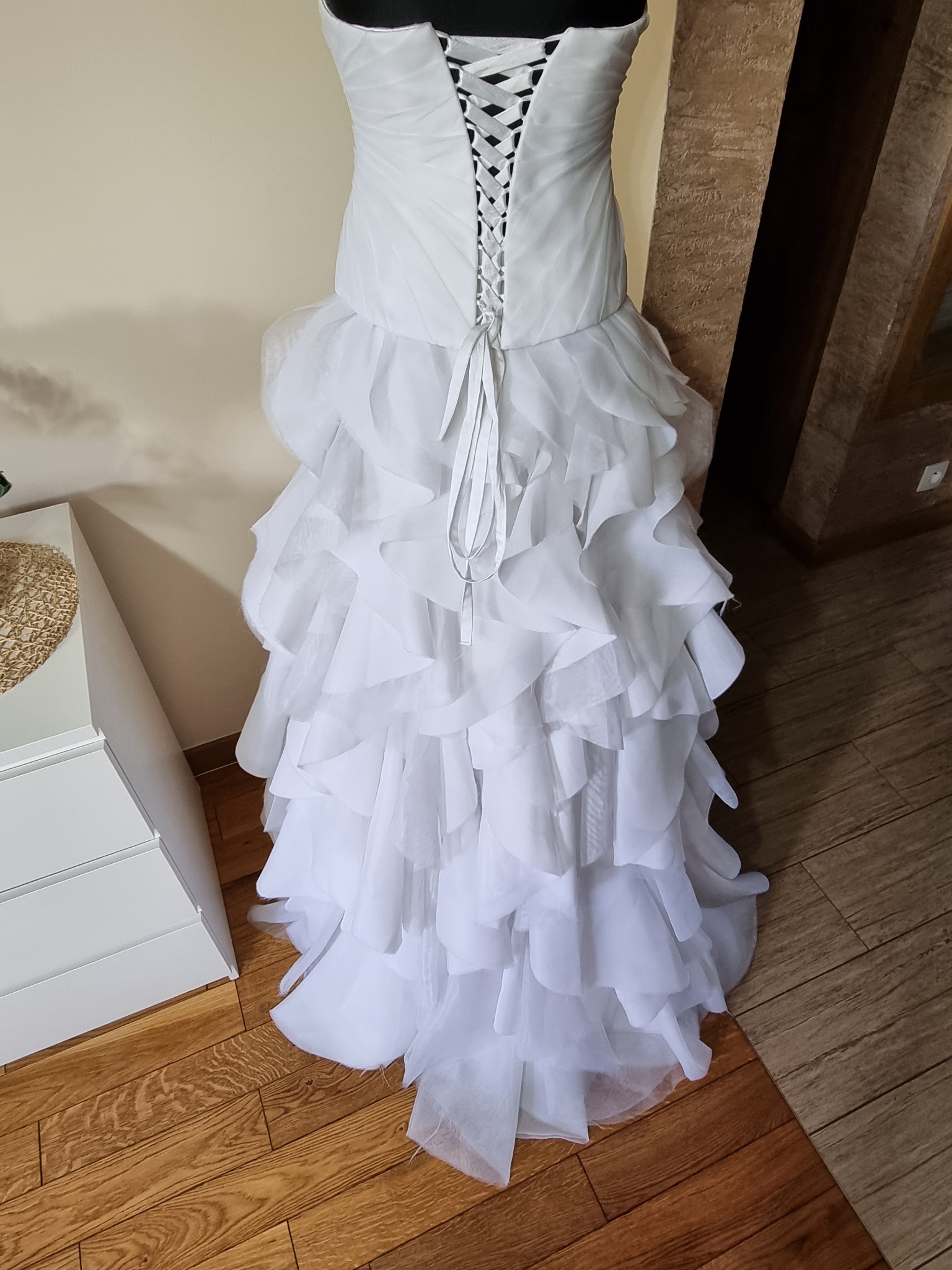 Tył białej sukni ślubnej z falbanami i sznurowaniem gorsetowym, widoczna na drewnianej podłodze obok białej komody.