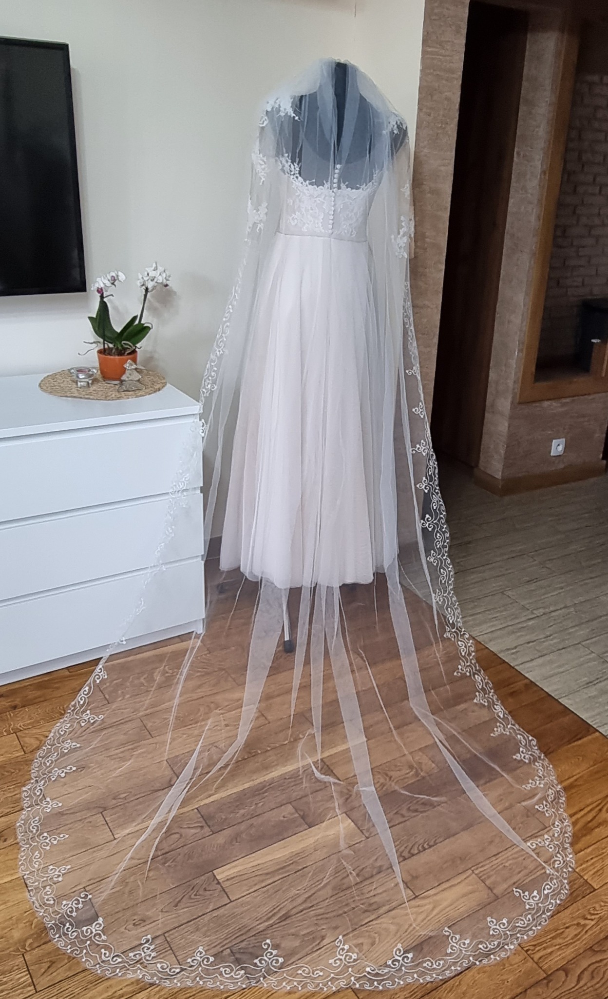 Elegancka suknia ślubna z długim, zdobionym welonem prezentowana na manekinie w salonie, obok komody z kwiatami.