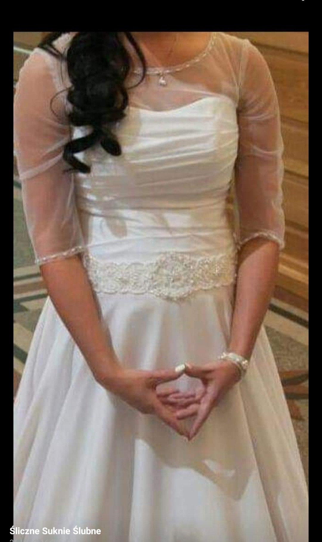 Panna młoda w białej sukni ślubnej z tiulowymi rękawami i ozdobnym pasem, dłonie złożone w charakterystyczny sposób, widoczny srebrny naszyjnik i bransoletka.