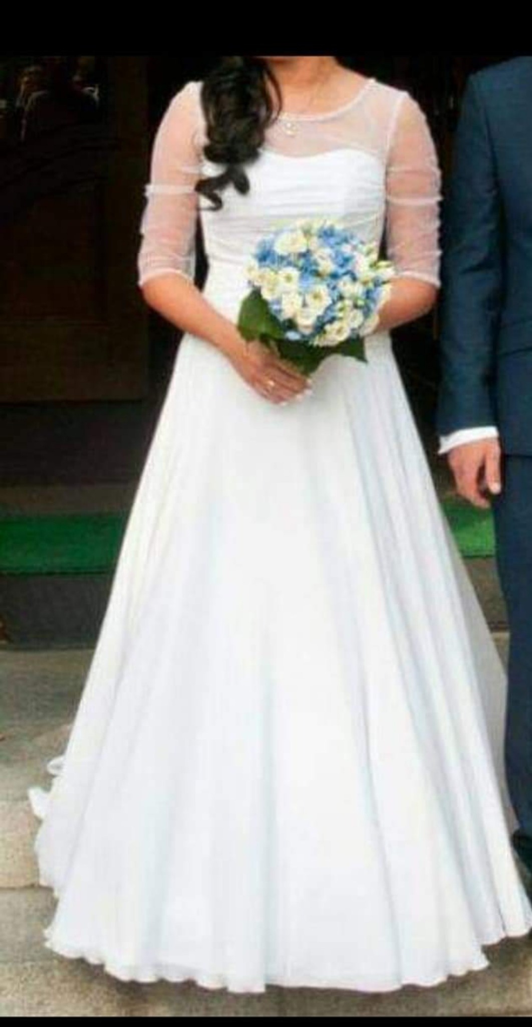 Panna młoda w białej sukni ślubnej z bukietem niebiesko-białych kwiatów, z półprzezroczystymi rękawami, stojąca obok mężczyzny w granatowym garniturze.