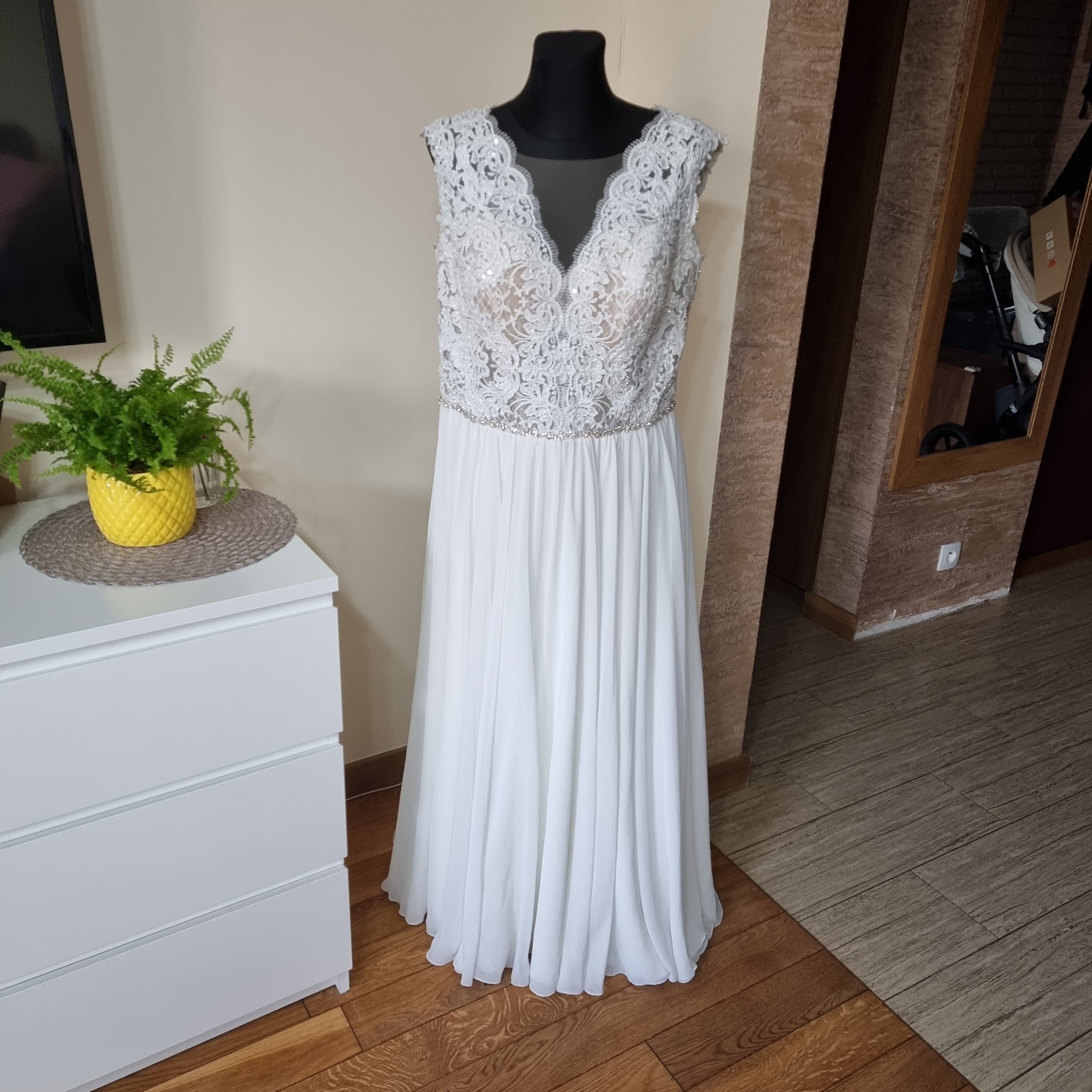 Elegancka suknia ślubna z koronkową górą i zwiewnym dołem, prezentowana na manekinie w domowym wnętrzu z komodą i rośliną w tle.