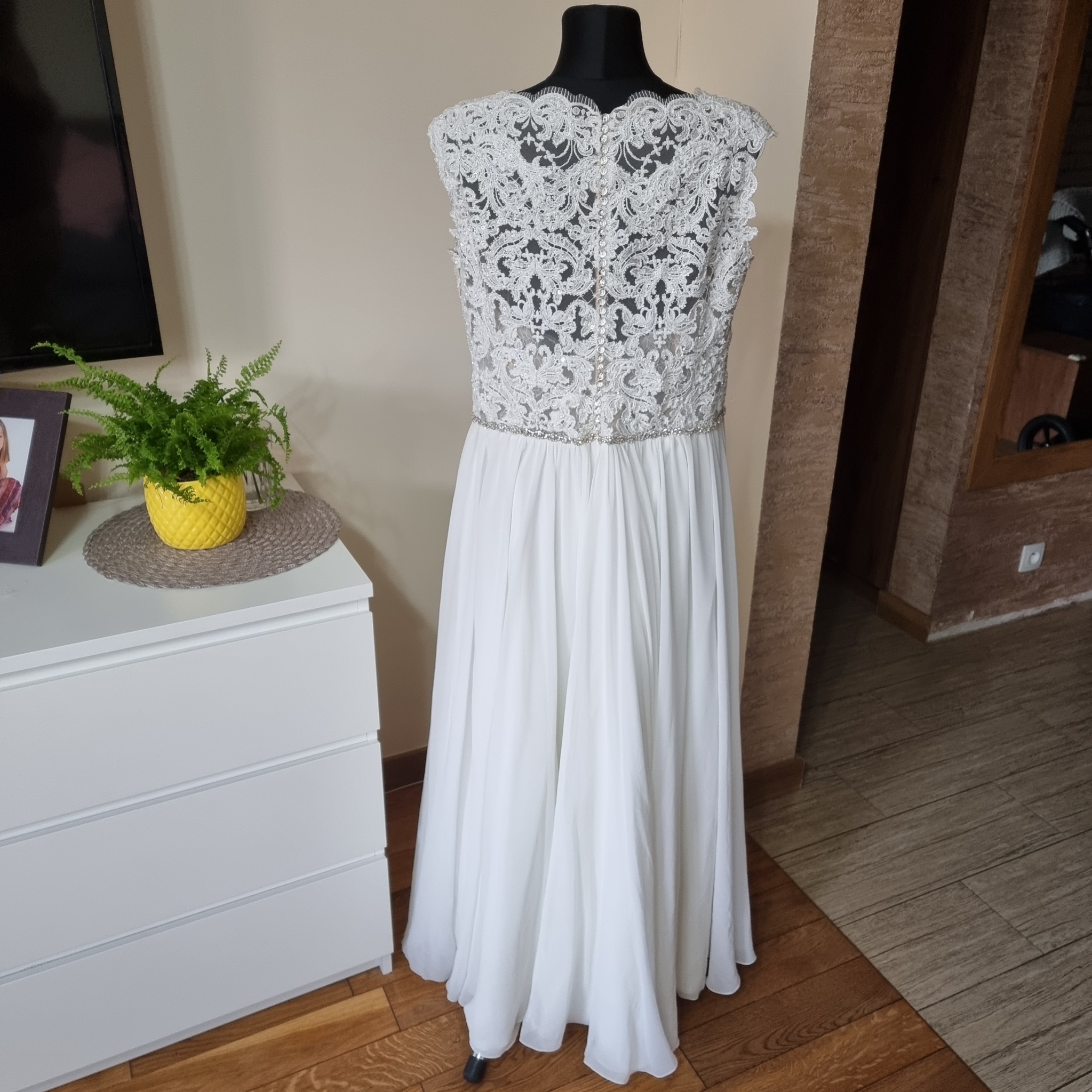 Elegancka suknia ślubna z koronkową górą i zwiewnym dołem, prezentowana na manekinie w domowym wnętrzu z komodą i kwiatami w tle.