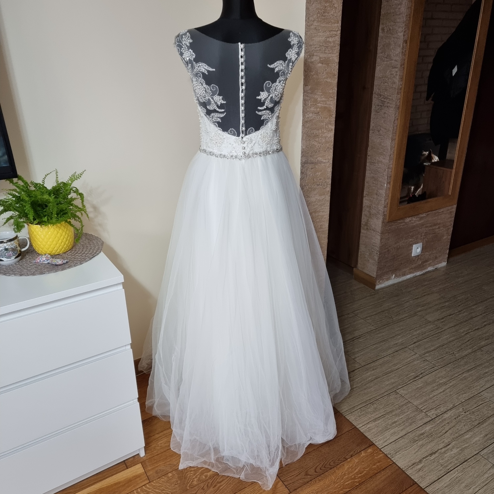 Suknia ślubna z koronkowym wykończeniem pleców, prezentowana na manekinie w pomieszczeniu z drewnianą podłogą i komodą z paprotką w tle.