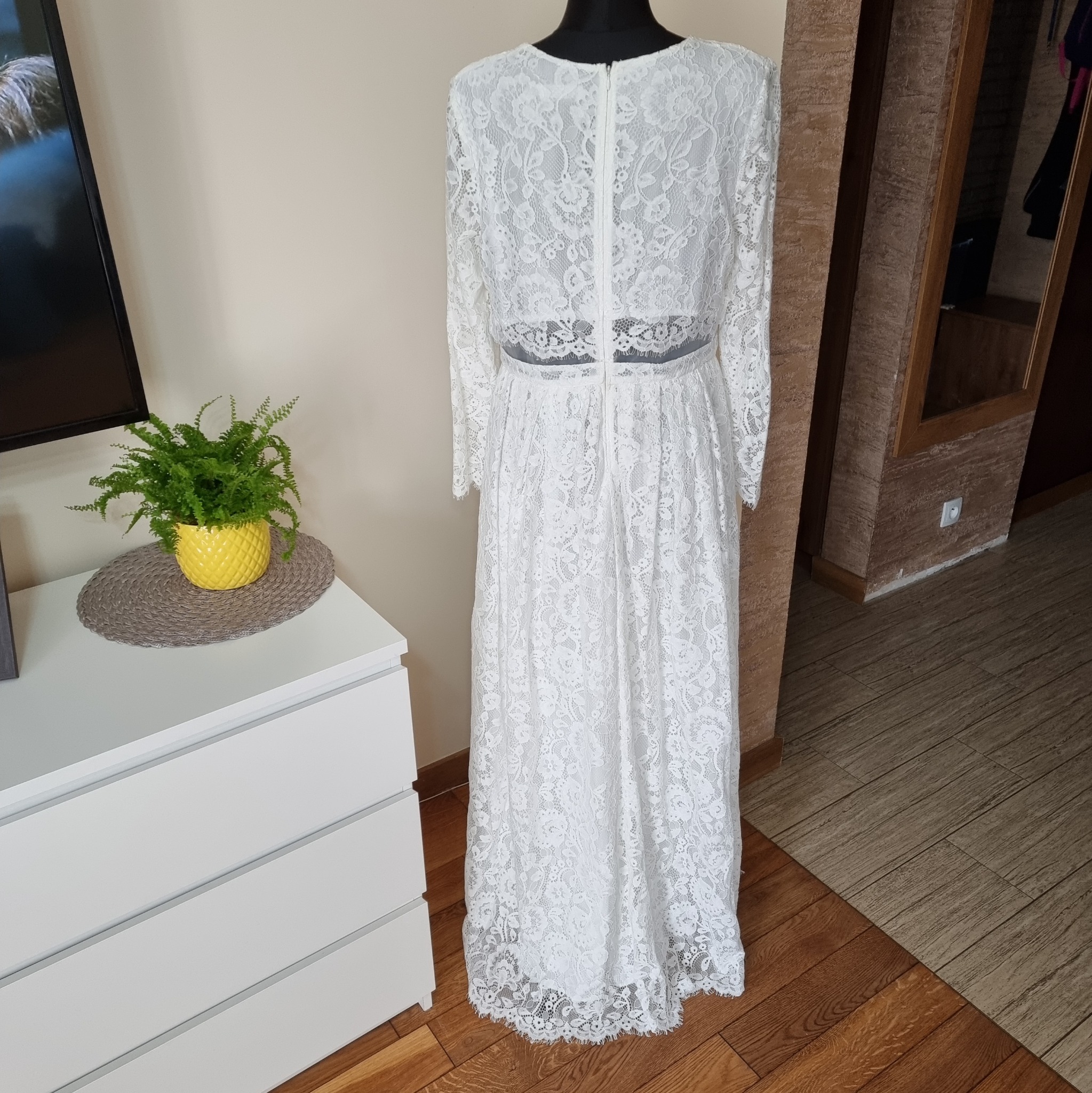 Biała koronkowa suknia z długim rękawem na manekinie, widok z tyłu, z suwakiem na plecach, obok komoda z paprotką w żółtej doniczce.