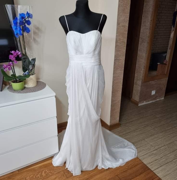 Elegancka suknia ślubna z drapowanym dołem na czarnym manekinie, obok komody z kwiatami.