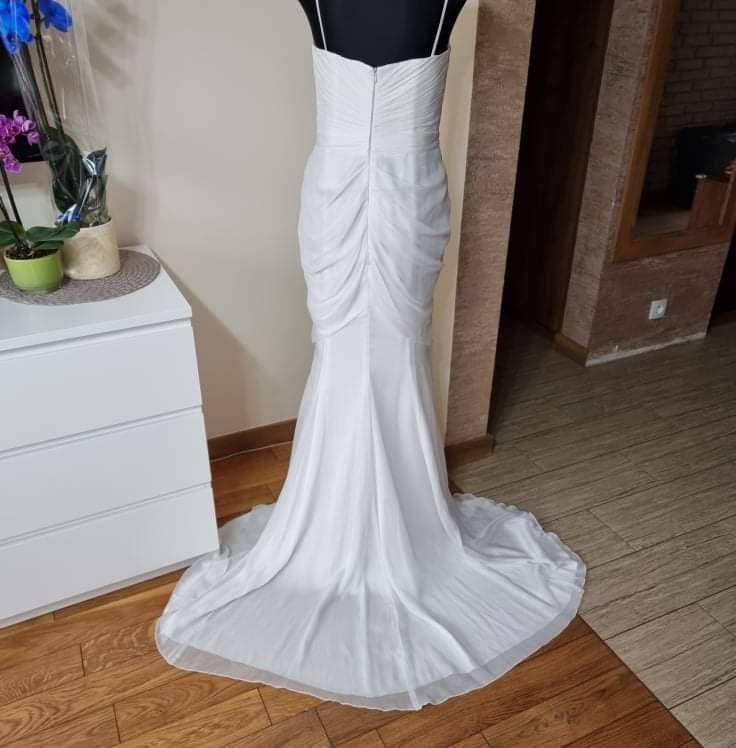 Elegancka suknia ślubna w kolorze białym, z drapowaniem na plecach i trenem, prezentowana na manekinie w pomieszczeniu z komodą i kwiatami.