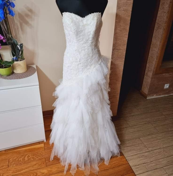 Koronkowa suknia ślubna typu rybka z falbanami na manekinie, obok biała komoda i kwiaty w doniczce.