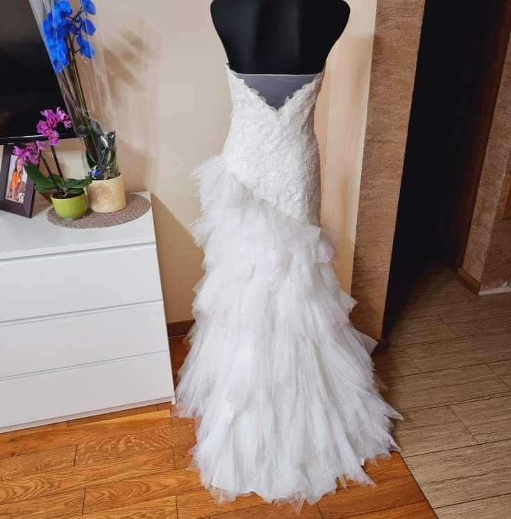 Koronkowa suknia ślubna z asymetrycznym, warstwowym trenem z tiulu, prezentowana na manekinie w domowym wnętrzu z kwiatami i komodą w tle.