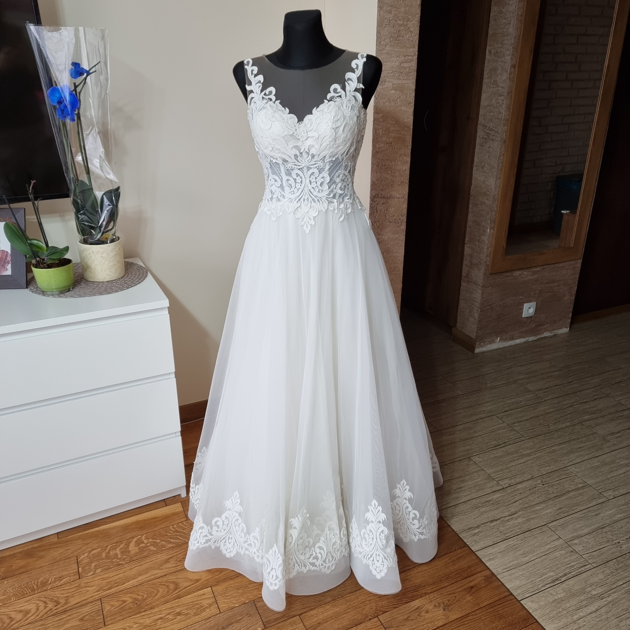 Elegancka suknia ślubna z koronkowym gorsetem i tiulowym dołem, prezentowana na manekinie w domowym wnętrzu z komodą i kwiatami w tle.