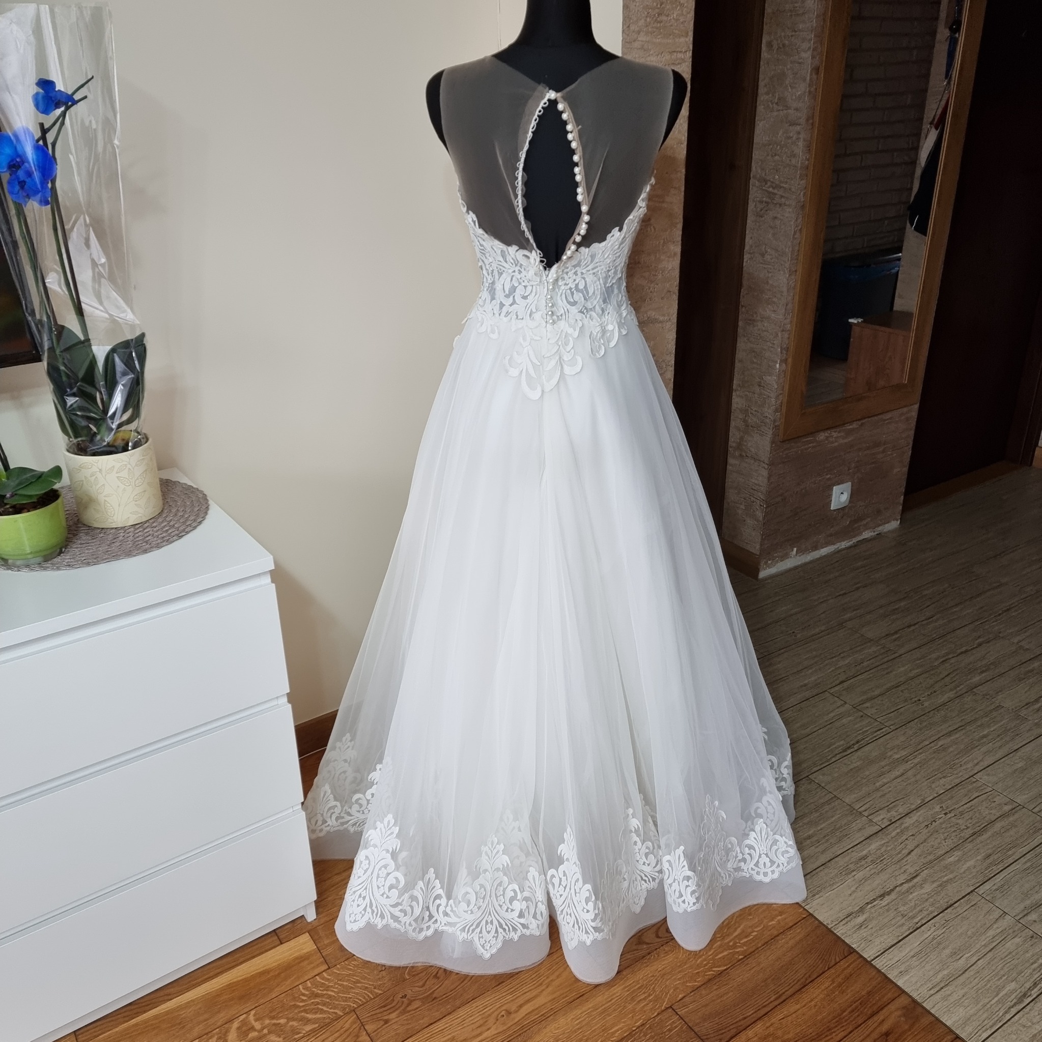 Tył białej sukni ślubnej z koronkowymi zdobieniami, perłowymi guzikami i tiulowym dekoltem na manekinie, obok komody z kwiatami.