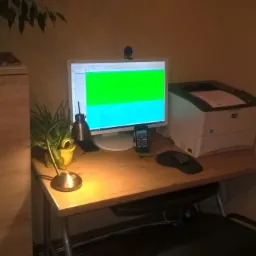 Biurko z komputerem, drukarką i lampką, na ekranie komputera widoczne dane w kolorach zielonym i niebieskim, obok kalkulator i myszka, całość w ciepłym oświetleniu