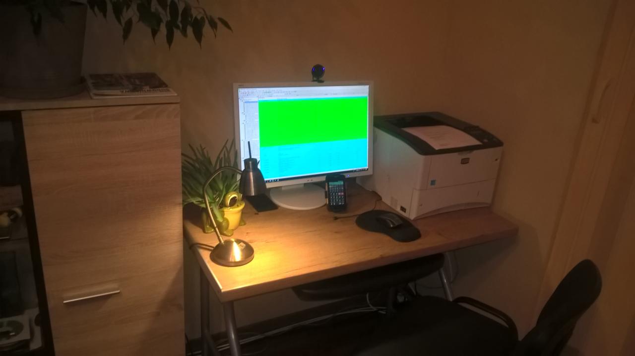 Biurko z komputerem, drukarką i lampką, na ekranie komputera widoczne dane w kolorach zielonym i niebieskim, obok kalkulator i myszka, całość w ciepłym oświetleniu