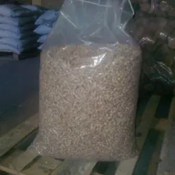 Wypełniony pelletem przezroczysty worek foliowy, ustawiony na drewnianej palecie, z widocznymi stosami worków w tle.