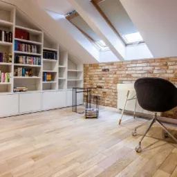 Biała zabudowa biblioteczna pod skosem dachu, drewniana podłoga, biurko z krzesłem na kółkach i ściana z cegły w domowym biurze.
