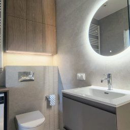 Baltic Development - Nowoczesna łazienka z szarymi płytkami, podświetlanym lustrem, wiszącą toaletą i drewnianą szafką. Minimalistyczny design, oświetlenie LED, chromowana armatura.