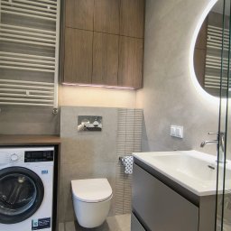 Baltic Development - Nowoczesna łazienka w szarościach z drewnianymi akcentami: podświetlana szafka nad toaletą, okrągłe lustro z ledowym podświetleniem, pralka obok grzejnika.