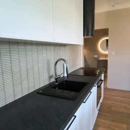 Baltic Development - Nowoczesna kuchnia z czarnym blatem i białymi szafkami, minimalistyczny design. W tle widoczna łazienka z okrągłym lustrem z podświetleniem LED.