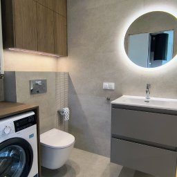 Baltic Development - Nowoczesna łazienka z szarą armaturą, podświetlanym lustrem i drewnianymi szafkami. Uwagę zwraca pralka wkomponowana w zabudowę, obok toalety.