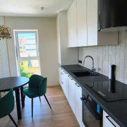 Baltic Development - Nowoczesna kuchnia z białymi szafkami, czarnym blatem i okrągłym stołem z zielonymi krzesłami. Widok na zewnątrz przez okno.