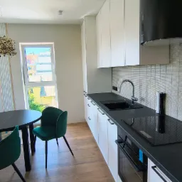 Nowoczesna kuchnia z białymi szafkami, czarnym blatem i okrągłym stołem z zielonymi krzesłami. Widok na zewnątrz przez okno.