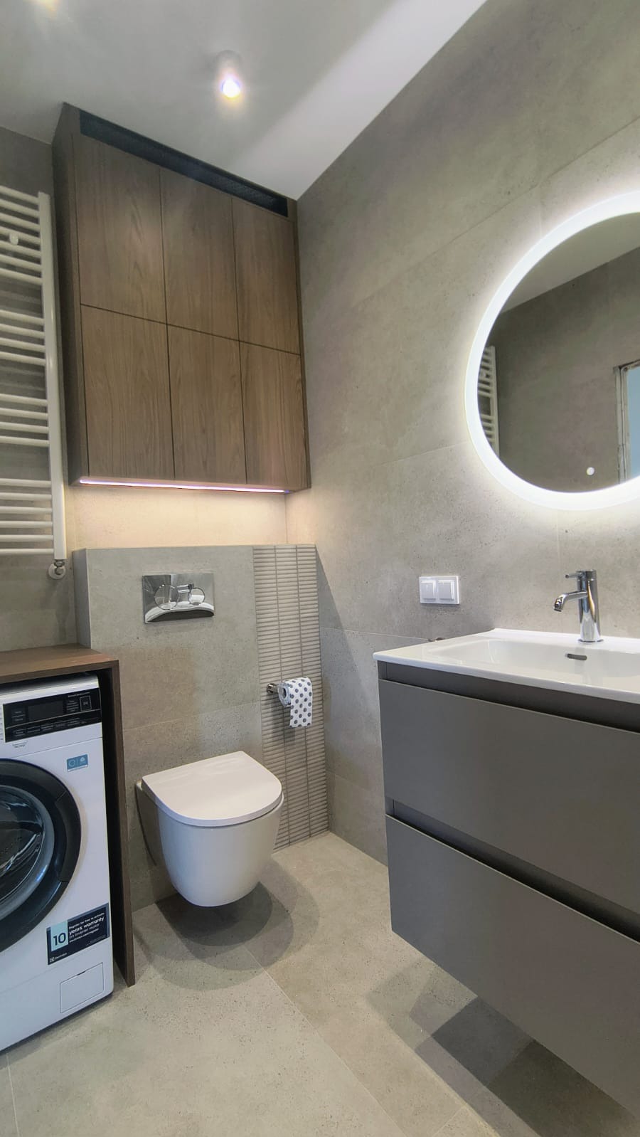 Nowoczesna łazienka w szarościach z podświetlanym lustrem, wiszącą toaletą i pralką obok grzejnika. Szafki nad toaletą z podświetleniem LED.