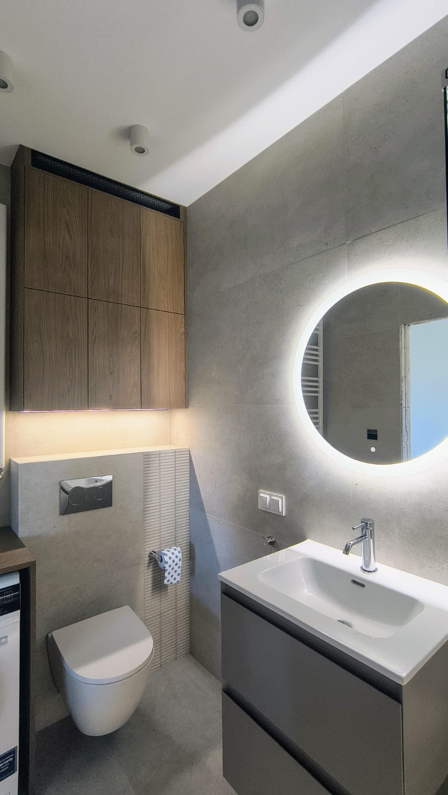 Nowoczesna łazienka w szarościach: podświetlane szafki nad toaletą, okrągłe lustro z podświetleniem LED, umywalka z szafką. Minimalistyczny design i stonowana kolorystyka.