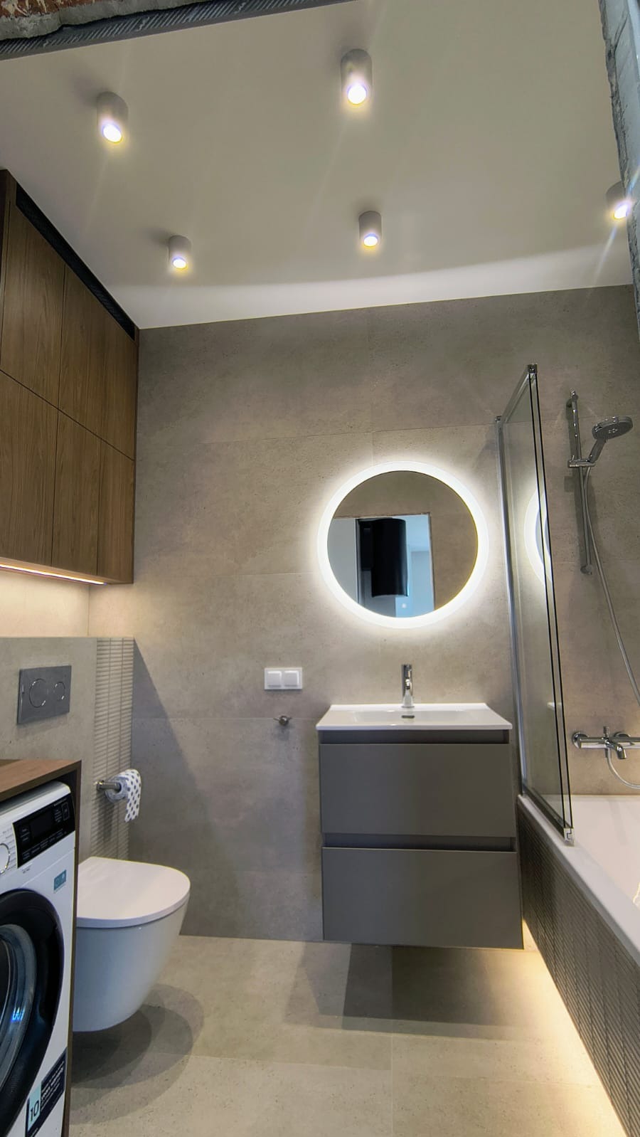 Nowoczesna łazienka z okrągłym lustrem LED, szarą szafką pod umywalką i pralką po lewej stronie. Minimalistyczny design, neutralna kolorystyka.
