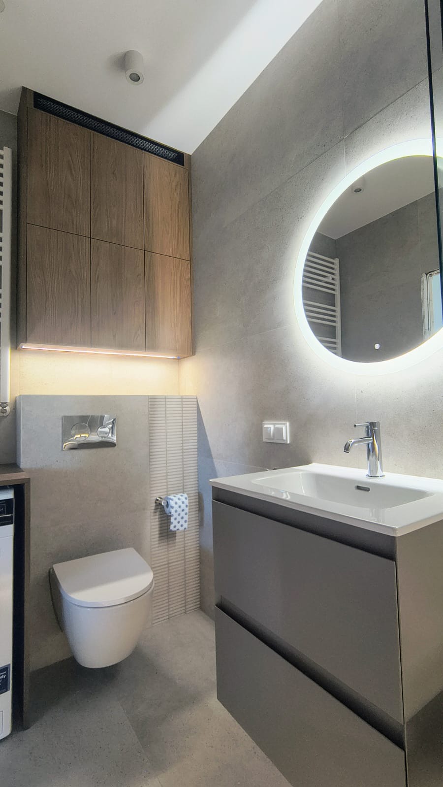 Nowoczesna łazienka z szarymi płytkami, podświetlanym lustrem, wiszącą toaletą i drewnianą szafką. Minimalistyczny design, oświetlenie LED, chromowana armatura.