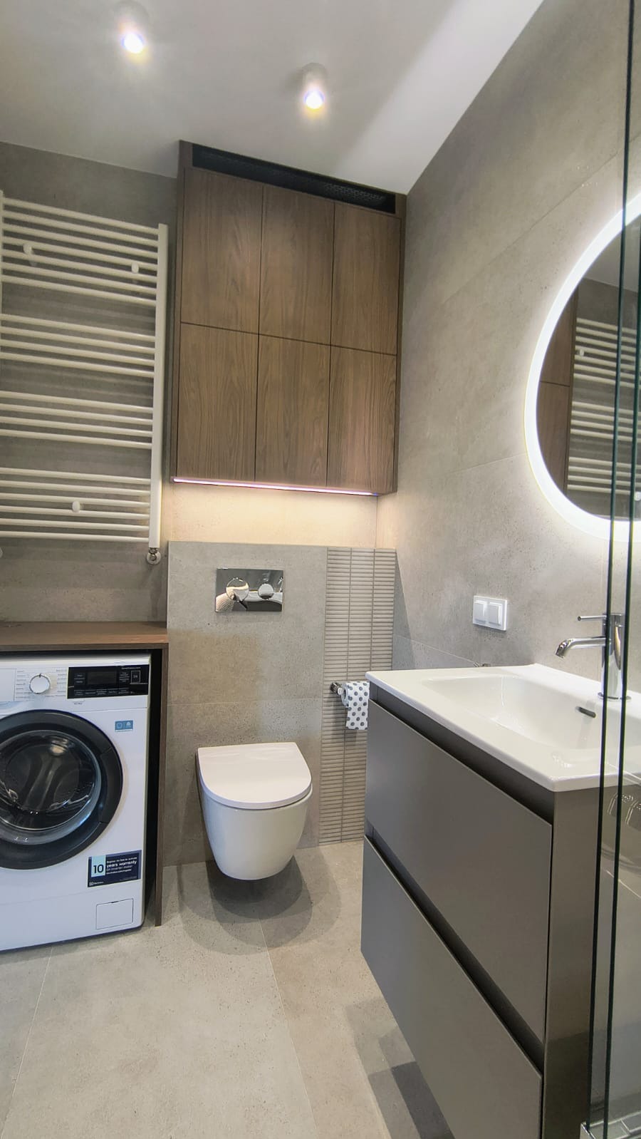 Nowoczesna łazienka w szarościach z drewnianymi akcentami: podświetlana szafka nad toaletą, okrągłe lustro z ledowym podświetleniem, pralka obok grzejnika.