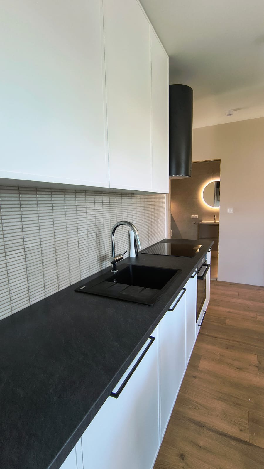 Nowoczesna kuchnia z czarnym blatem i białymi szafkami, minimalistyczny design. W tle widoczna łazienka z okrągłym lustrem z podświetleniem LED.