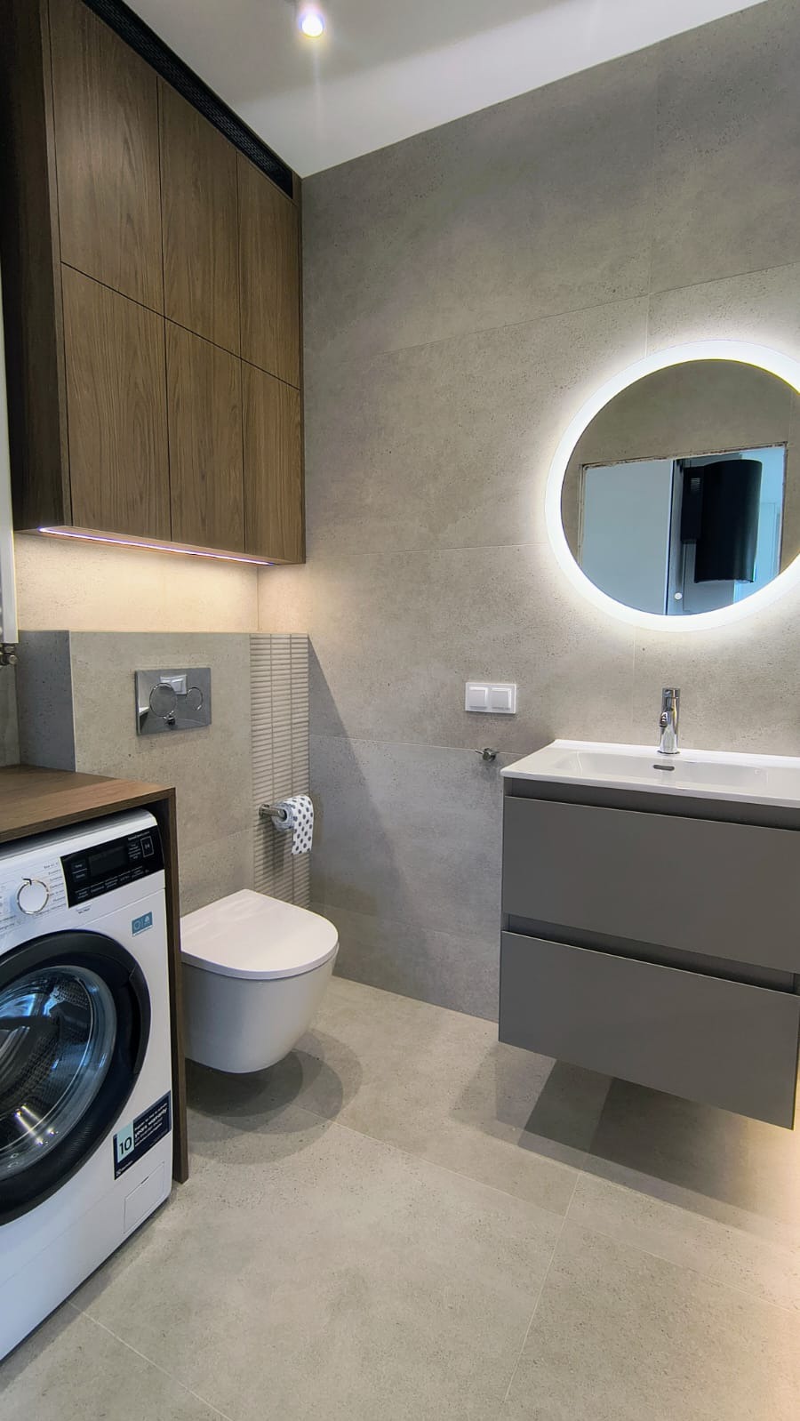 Nowoczesna łazienka z szarą armaturą, podświetlanym lustrem i drewnianymi szafkami. Uwagę zwraca pralka wkomponowana w zabudowę, obok toalety.
