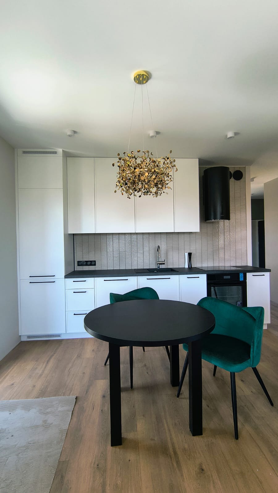 Nowoczesna kuchnia z białymi szafkami, czarnym blatem, okrągłym stołem i zielonymi krzesłami. Nad stołem designerska złota lampa.