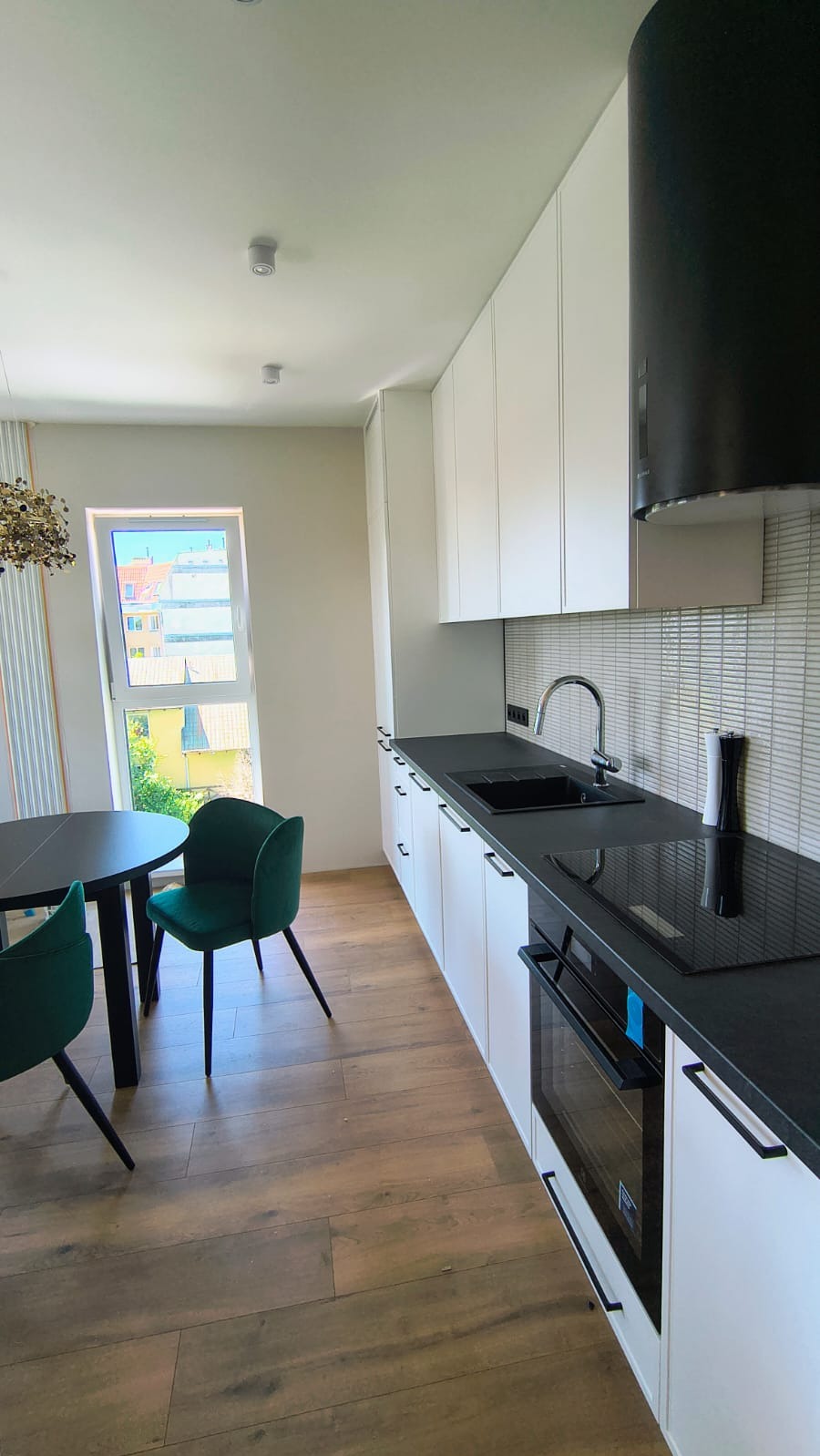 Nowoczesna kuchnia z białymi szafkami, czarnym blatem i okrągłym stołem z zielonymi krzesłami. Widok na zewnątrz przez okno.