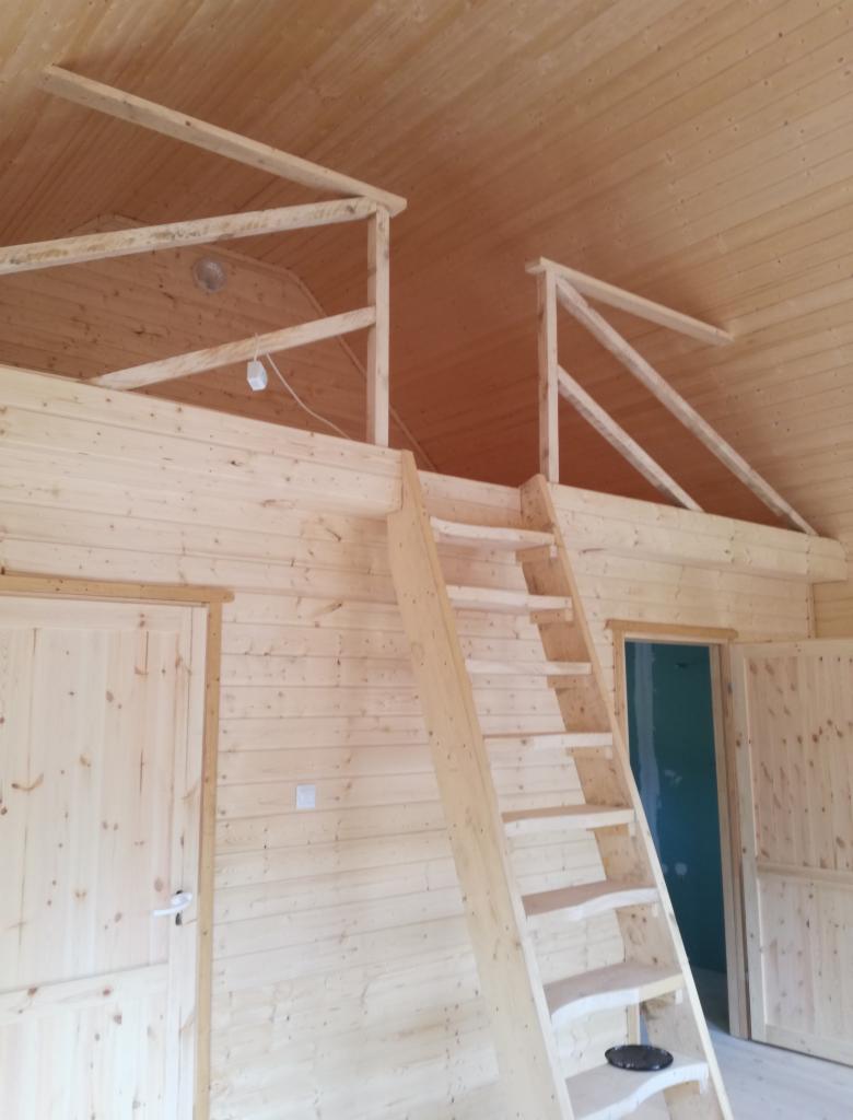 Wnętrze drewnianego domu z antresolą, widoczne schody drabinkowe, drewniane ściany i drzwi, surowy stan wykończenia.