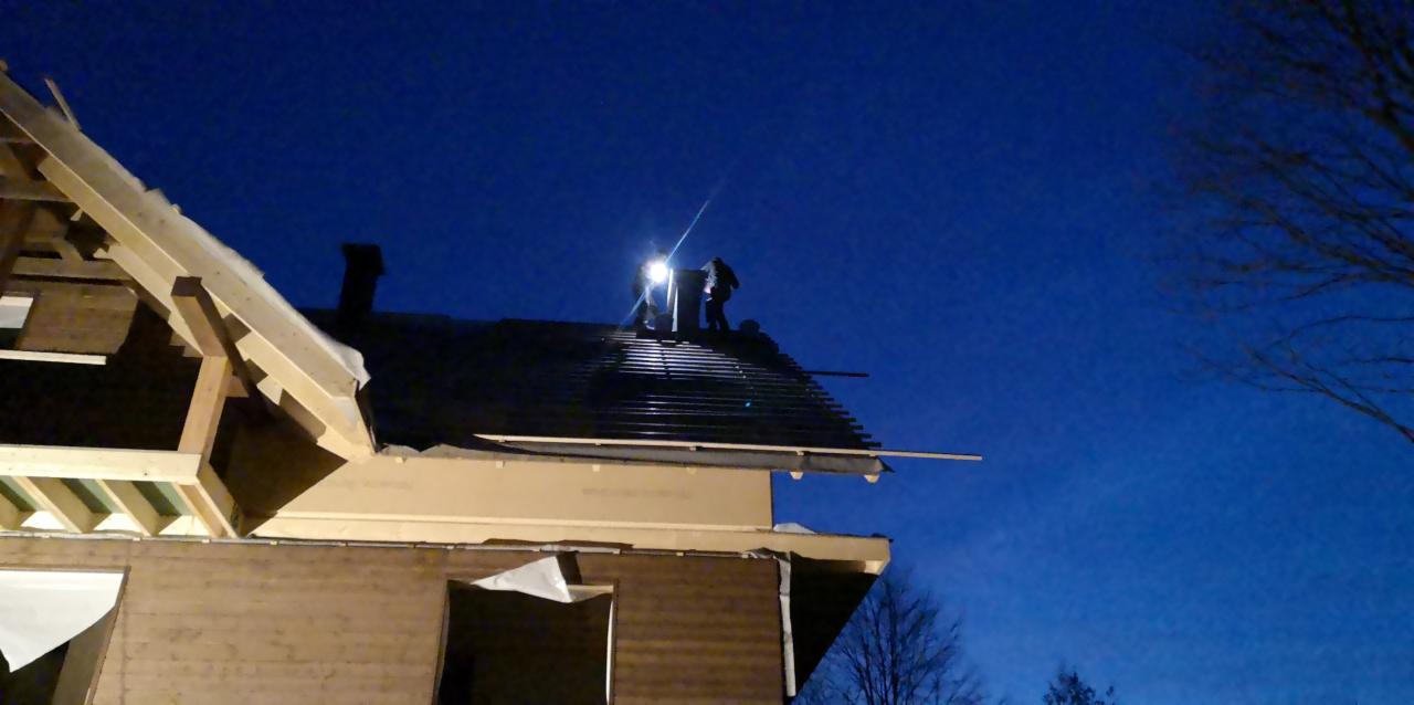 Budowa domu w nocy: ekipa montuje dach pod reflektorami, widoczna konstrukcja szkieletowa i fragment elewacji.