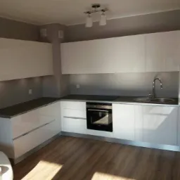 Nowoczesna, minimalistyczna kuchnia z białymi, lakierowanymi frontami, szarym blatem i drewnopodobną podłogą. Widoczne subtelne oświetlenie podszafkowe.