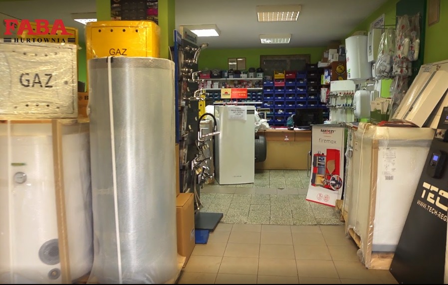 Wnętrze hurtowni instalacyjnej z widokiem na kotły gazowe i zbiorniki na wodę, niektóre jeszcze zapakowane w folię ochronną, z logo hurtowni widocznym w lewym górnym rogu.