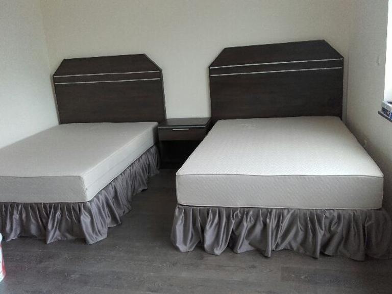 Dwa łóżka pojedyncze z ciemnobrązowymi wezgłowiami z jasnymi paskami i szarymi falbanami, stojące obok stolika nocnego w pokoju o jasnych ścianach.