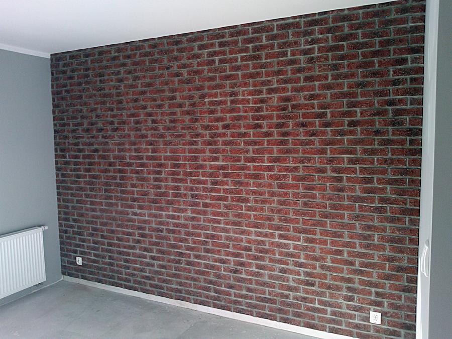Ściana wykończona czerwoną cegłą klinkierową z szarą fugą, widoczny fragment grzejnika i gniazdka elektrycznego, szara ściana boczna, surowy beton na podłodze.