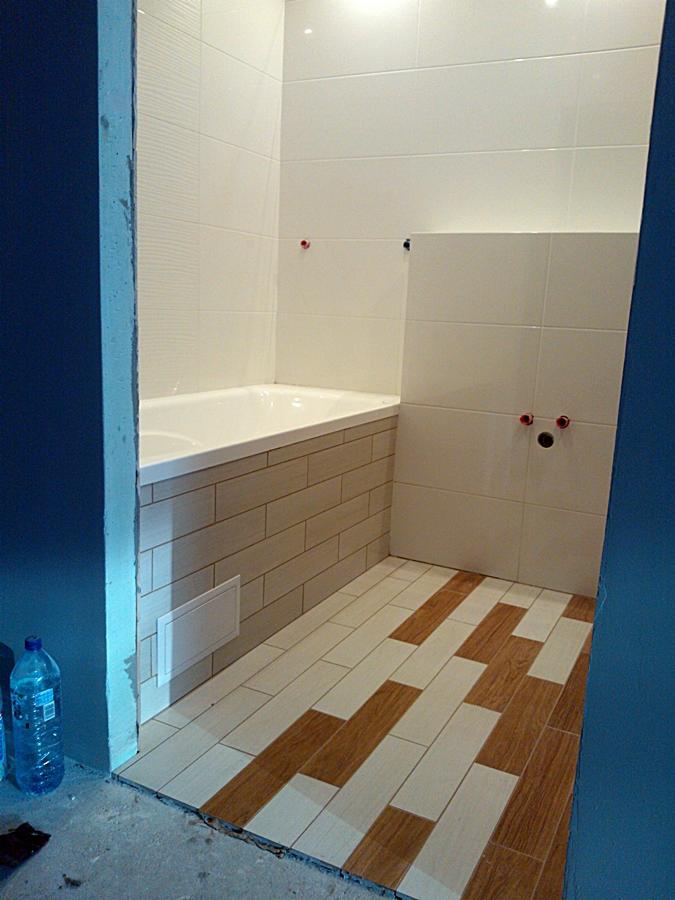 Wnętrze łazienki w trakcie prac wykończeniowych: ułożone płytki podłogowe w odcieniach brązu i beżu, obudowa wanny wykończona płytkami imitującymi cegłę, białe płytki ścienne, widoczne przyłącza...