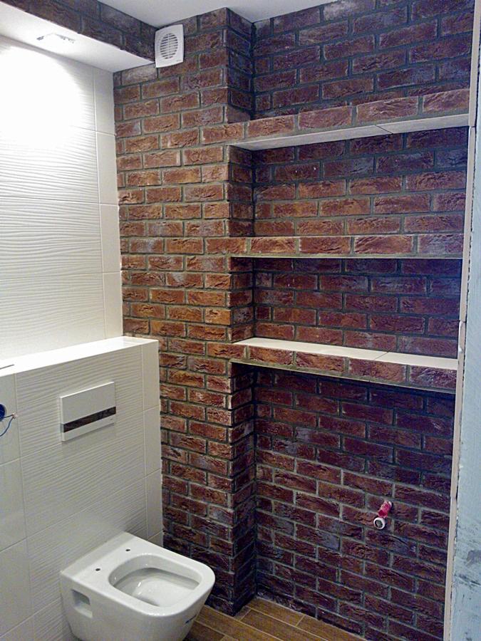 Łazienka w trakcie remontu: toaleta podwieszana z białymi płytkami strukturalnymi, ściana z cegły klinkierowej z wnękami na półki, wentylator sufitowy.