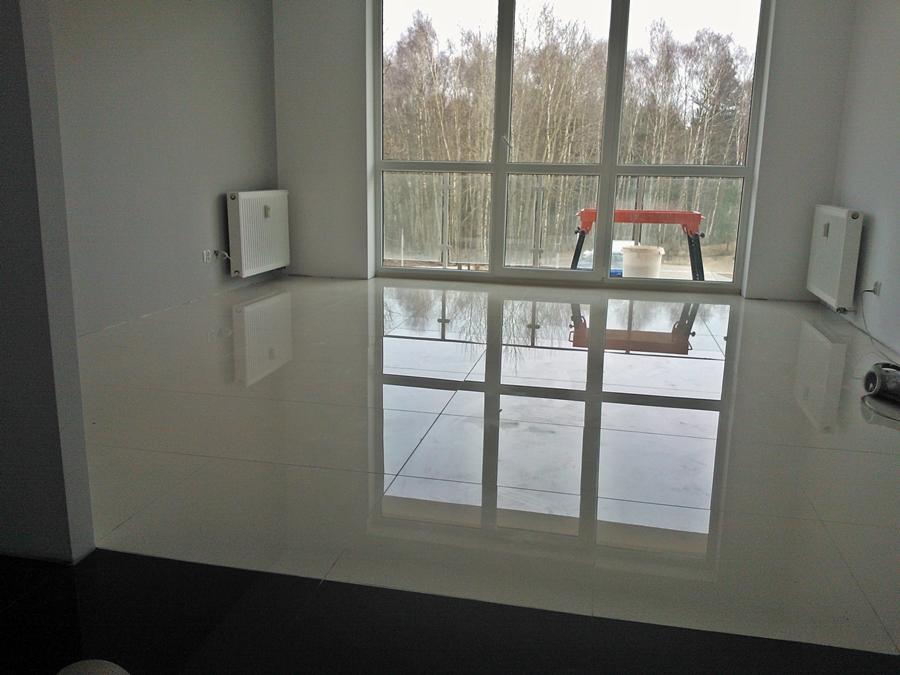 Błyszcząca, jasna podłoga z płytek w nowym, pustym pokoju z dużym oknem, przez które widać drzewa. Widoczny grzejnik na ścianie i kozioł malarski z wiadrem w tle.