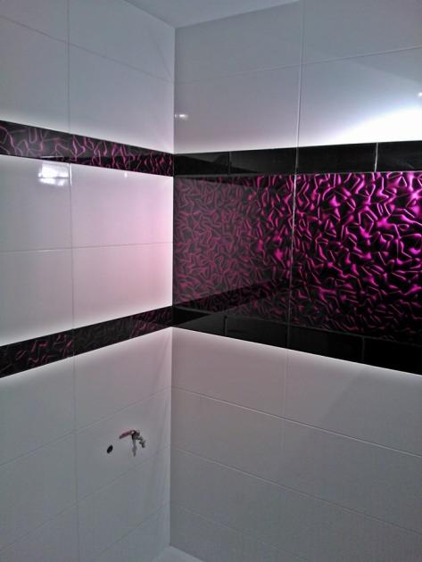 Wykończona ściana łazienki z białymi płytkami i dekoracyjnym pasem z czarnych płytek z fioletowym wzorem, oświetlenie LED wzdłuż pasów, widoczny fragment instalacji wodnej.