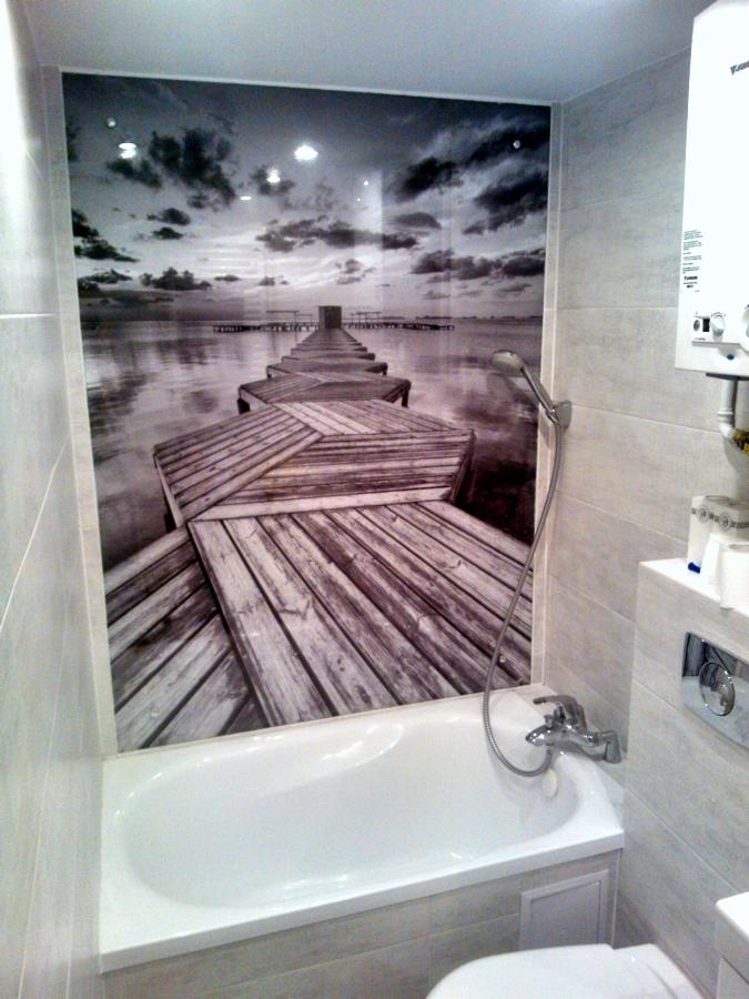 Biała wanna w łazience z fototapetą przedstawiającą drewniane molo i zachmurzone niebo nad wodą, obok grzejnik i toaleta