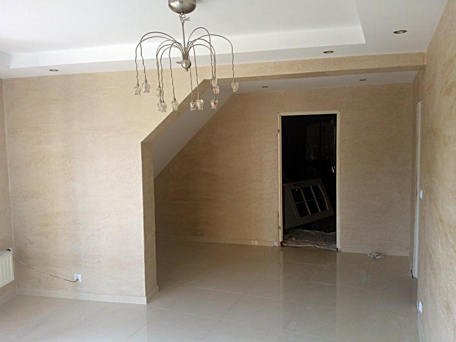 Pusta przestrzeń w odcieniach beżu z błyszczącą podłogą z płytek, dekoracyjną lampą sufitową i ścianami z efektem trawertynu, z widocznym oknem w głębi niedokończonego pomieszczenia.