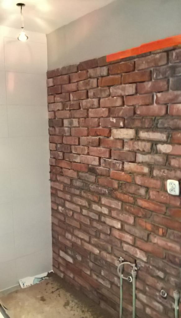 Ściana łazienki wyłożona czerwoną cegłą, widoczne przyłącza wodne, gniazdko elektryczne i fragment podłogi w trakcie prac wykończeniowych.