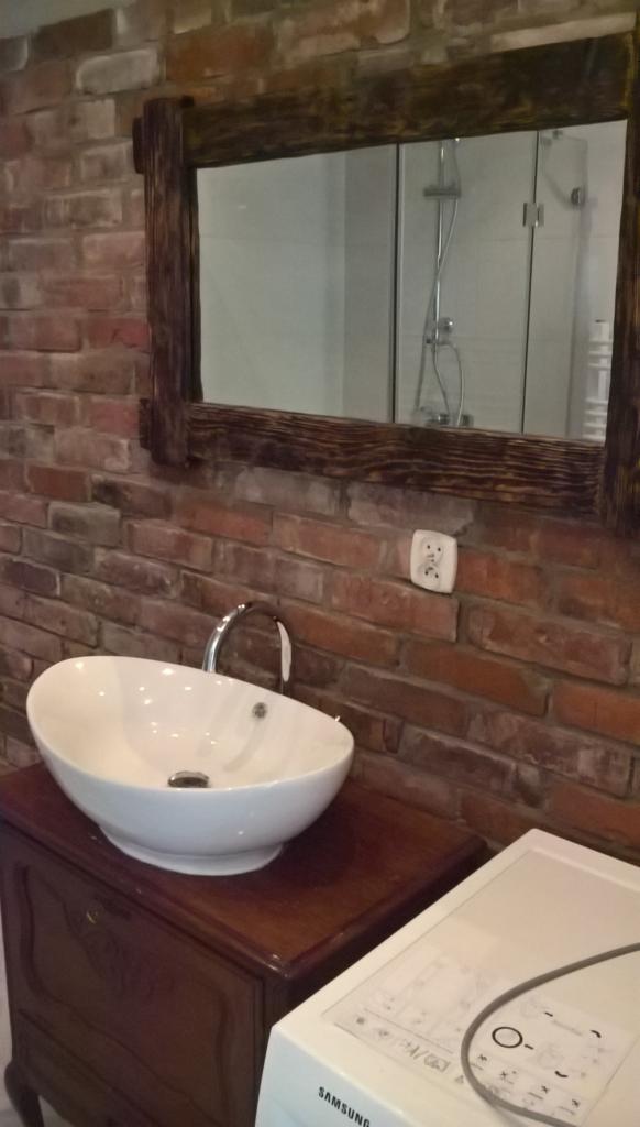 Umywalka nablatowa w stylu retro na drewnianej szafce, z kranem w kolorze chromu, na tle ściany z czerwonej cegły i lustra w drewnianej ramie, w którym odbija się kabina prysznicowa.
