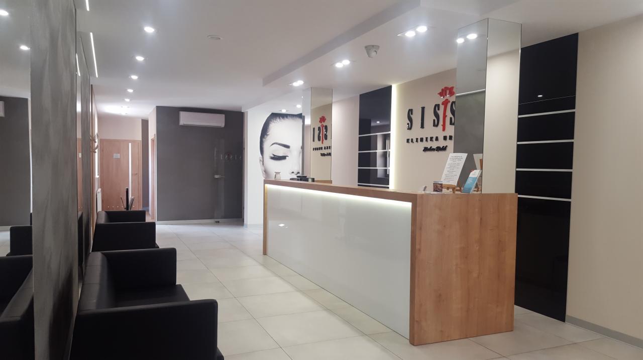 Jasne, nowoczesne wnętrze kliniki z recepcją, logo 'SISIS' i grafiką twarzy kobiety na ścianie, czarne sofy w poczekalni.