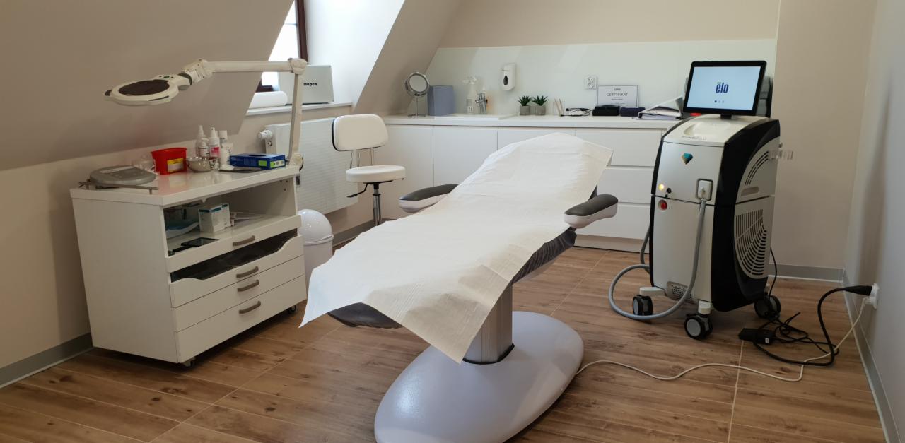 Jasne wnętrze gabinetu z fotelem kosmetycznym pokrytym białym papierem, obok widoczny laser kosmetyczny z ekranem dotykowym, białe meble i drewniana podłoga.